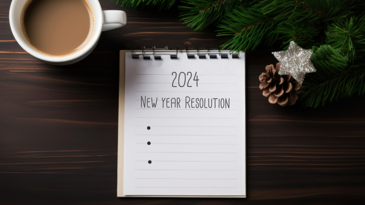 A notepad has 2024 listed as a header with bullet points waiting for New Year Resolutions to be filled in. The notepad is on a desk with coffee.